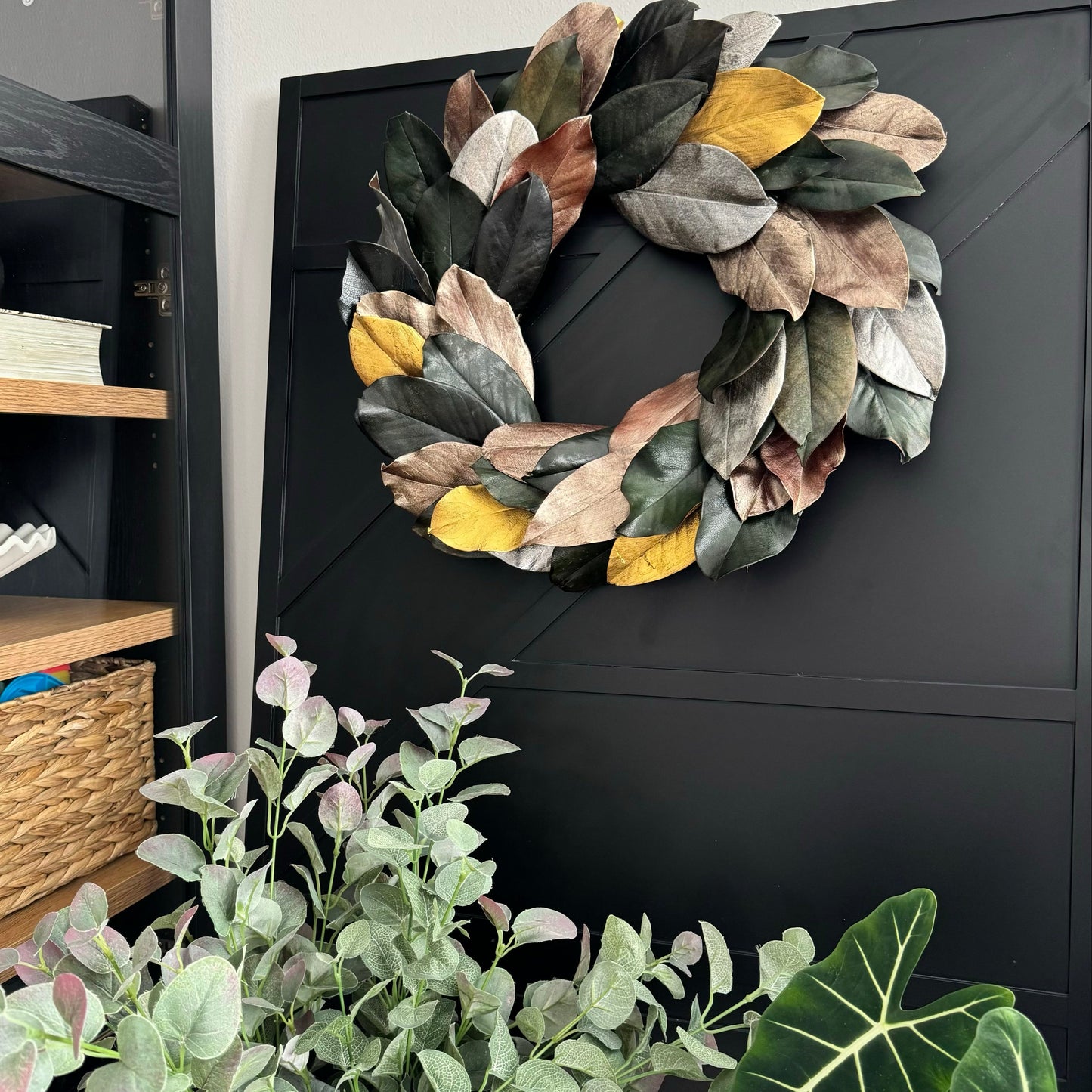 preserved gold and silver magnolia wreath hanging on a black patterned wooden panel with greenery in the front