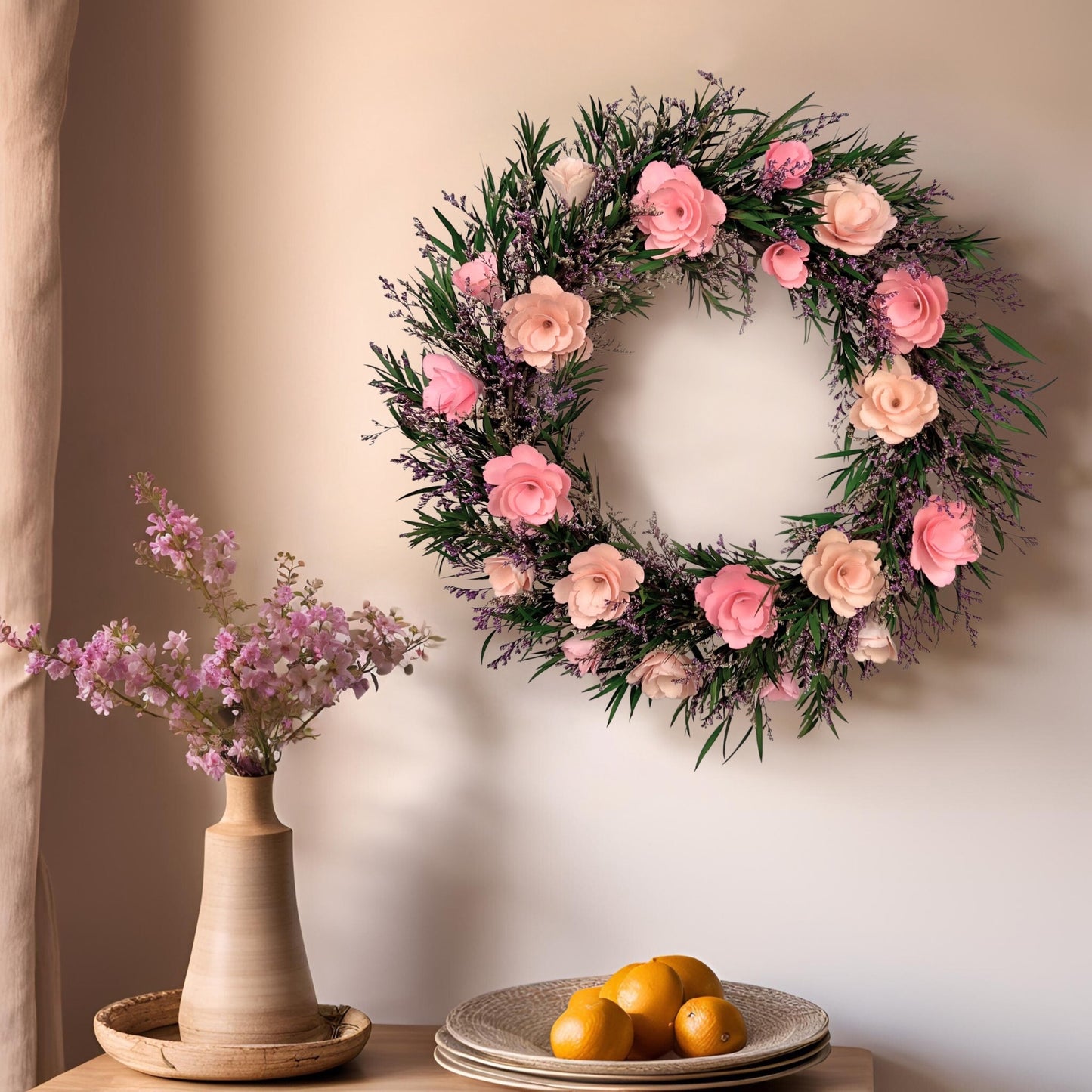 Floral wreath with pink flowers on a wall, next to a vase with pink flowers and a plate of oranges.