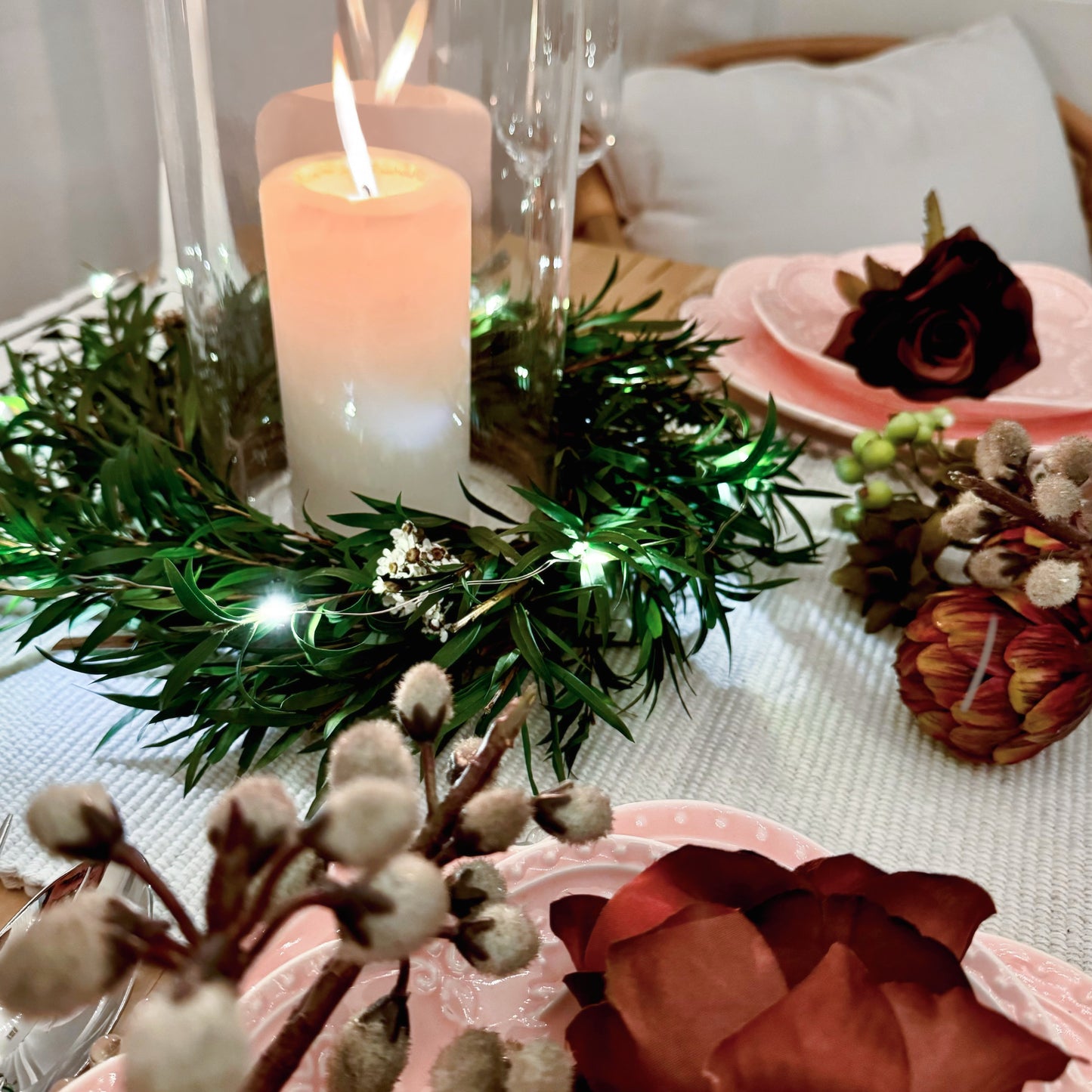 Festive holiday table setting with preserved greenery candle ring wreath, glass hurricane candle holder, and warm string lights surrounded by floral dinnerware