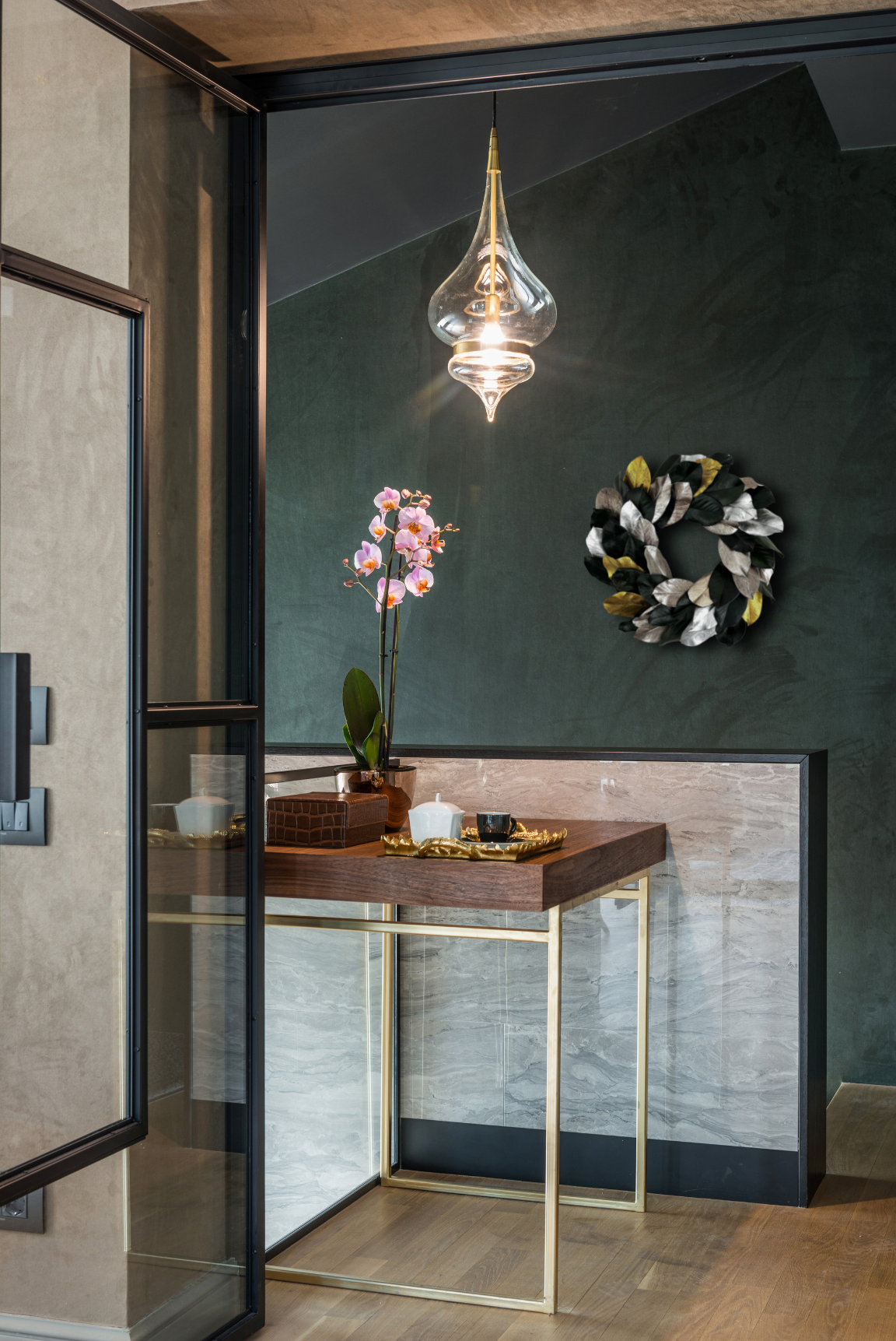 Overhead view of modern entryway featuring a preserved magnolia leaf wreath on a dark green wall, glass pendant light, and wood-accented console with orchid plant